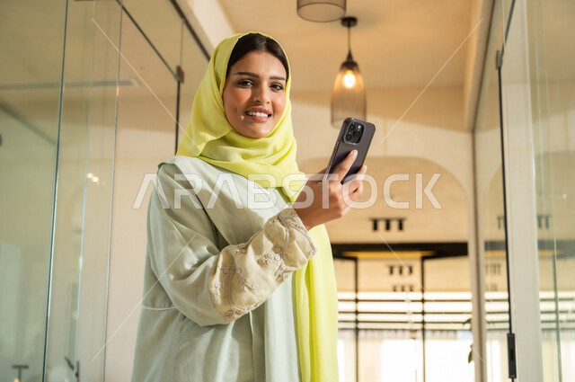 Looking at the camera with gestures of joy and pleasure, using modern devices and technologies to accomplish tasks, using mobile phone applications and services, a young Saudi Arabian Gulf woman wearing an abaya and hijab standing inside the company headquarters holding a mobile phone and reading a message, office jobs and professions for women