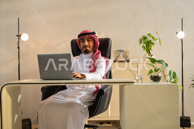 النظر الى الكاميرا بايماءات الثقة بالنفس، بيئة عمل مريحة في الشركات السعودية، عقد اجتماع عمل عبر الانترنت, رجل عربي خليجي سعودي يرتدي الثوب التقليدي والشماغ يقوم بتصفح مواقع أخبار السوشال ميديا عبر جهاز الحاسوب, استخدام الأجهزة والتقنيات الحديثة والمتطورة