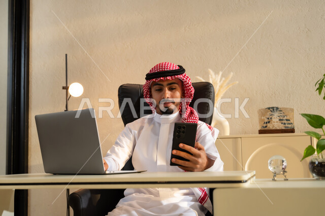 إيماءات البهجة والسرور، التسوق الالكتروني عبر برامج الهاتف المحمول، الدردشة مع الأهل والأصدقاء، رجل أعمال عربي سعودي خليجي مبتسم يرتدي الشماغ والثوب التقليدي يجلس على مكتب أمام اللابتوب يمسك الجوال ويقوم بالرد على رسالة ما، بيئة عمل مريحة في الشركات السعودية