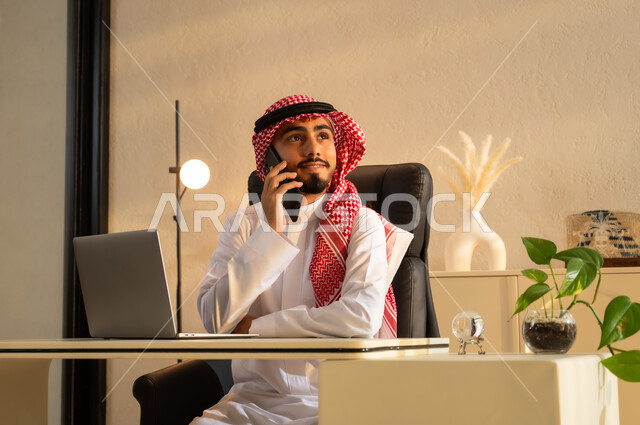 النظر الى شيء ما بإيماءات البهجة والسرور، استخدام جهاز اللابتوب والتقنيات الحديثة، شاب عربي سعودي خليجي مبتسم يرتدي الزي التقليدي يجلس على مكتب يقوم بإجراء مكالمة هاتفيه باستخدام الهاتف المحمول, مهن ووظائف مكتبية، بيئة عمل مريحة في الشركات السعودية