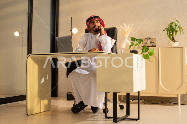مهن ووظائف ادارية، استخدام جهاز اللابتوب والتقنيات الحديثة، شاب عربي سعودي خليجي مبتسم يرتدي الزي التقليدي يجلس على مكتب يقوم بإجراء مكالمة هاتفيه باستخدام الهاتف المحمول ينظر الى شيء ما بإيماءات البهجة والسرور, بيئة عمل مريحة في الشركات السعودية