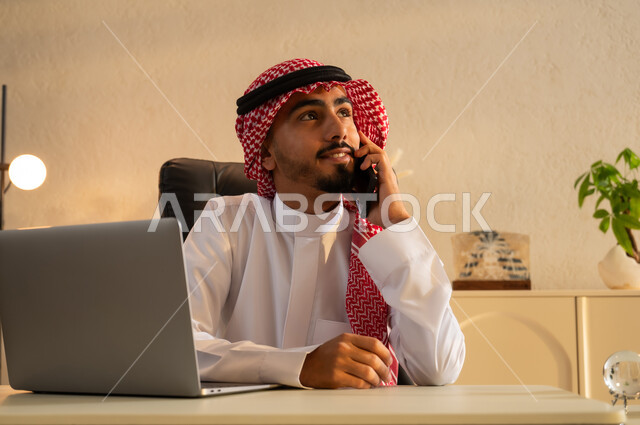 بيئة عمل مريحة في الشركات السعودية، مهن ووظائف ادارية، استخدام جهاز اللابتوب والتقنيات الحديثة، شاب عربي سعودي خليجي مبتسم يرتدي الزي التقليدي يجلس على مكتب يقوم بإجراء مكالمة هاتفيه باستخدام الهاتف المحمول ينظر الى شيء ما بإيماءات البهجة والسرور