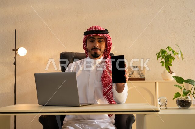 عرض بشاشة سوداء فارغة على الجوال, استعمال تطبيقات وخدمات الهاتف المحمول, شاب عربي خليجي سعودي يرتدي الثوب التقليدي والشماغ يجلس على مكتب أمام اللابتوب، استخدام الأجهزة والتقنيات الحديثة, التواصل مع الأهل والأصدقاء عبر الأونلاين