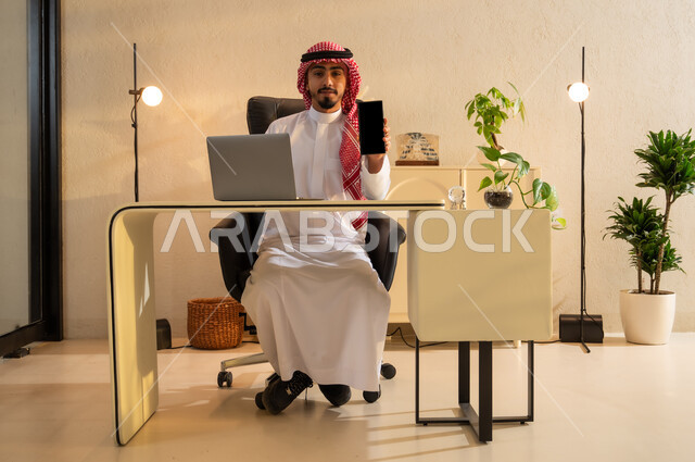 Using modern devices and technologies, displaying a blank black screen on a mobile phone, using mobile phone applications and services, a young Saudi Arabian Gulf woman wearing an abaya and hijab sitting at a desk in front of a laptop, communicating with family and friends online