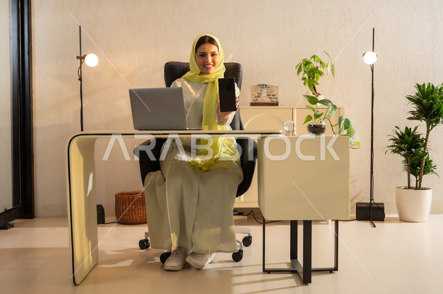عرض بشاشة سوداء فارغة على الجوال, استعمال تطبيقات وخدمات الهاتف المحمول, شابة عربية خليجية سعودية ترتدي العباية والحجاب تجلس على مكتب أمام اللابتوب، استخدام الأجهزة والتقنيات الحديثة, التواصل مع الأهل والأصدقاء عبر الأونلاين