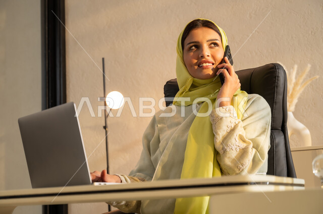 Using laptop and modern technology, looking at something with gestures of joy and pleasure, close-up of a young Saudi Arabian Gulf woman wearing abaya and hijab sitting at a desk making a phone call using a mobile phone, office jobs and professions, comfortable work environment in Saudi companies