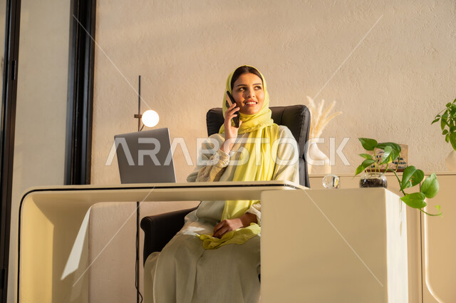 النظر الى شيء ما بإيماءات البهجة والسرور، استخدام جهاز اللابتوب والتقنيات الحديثة، شابة عربية سعودية خليجية ترتدي العباية والحجاب تجلس على مكتب تقوم بإجراء مكالمة هاتفيه باستخدام الهاتف المحمول, مهن ووظائف مكتبية، بيئة عمل مريحة في الشركات السعودية
