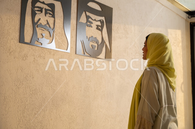 استذكار اليوم الوطني، مفهوم حب الوطن والانتماء اليه، صورة من الخلف لشابة عربية خليجية سعودية ترتدي العباية والحجاب تقف أمام لوحات فنية تحمل صور لملوك المملكة العربية السعودية، الفخر والاعتزاز بإنجازات البلاد