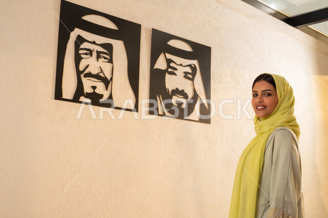 النظر الى الكاميرا بايماءات الفخر والاعتزاز بإنجازات البلاد، استذكار اليوم الوطني، مفهوم حب الوطن والانتماء اليه، شابة عربية خليجية سعودية ترتدي العباية والحجاب تقف أمام لوحات فنية تحمل صور لملوك المملكة العربية السعودية