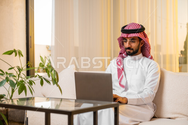 تصفح مواقع أخبار السوشال ميديا عبر جهاز الحاسوب، استخدام الأجهزة والتقنيات الحديثة والمتطورة، شاب عربي خليجي سعودي يجلس على كنبة مريحة يقوم بالتسوق عبر الانترنت