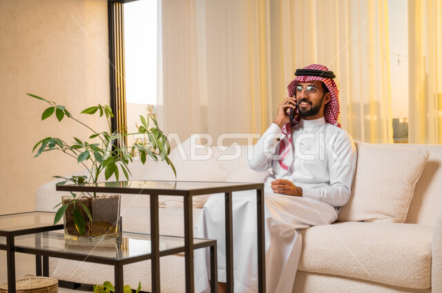 تعابير وإيماءات البهجة والسعادة، التواصل مع الأصدقاء والاقارب باستخدام الجوال، الرد على الاتصالات والمكالمات، سماع أخبار سعيدة عبر الهاتف المحمول، بورتريه لشاب عربي خليجي سعودي يرتدي الزي التقليدي يجلس على كنبة مريحة يقوم بإجراء مكالمة هاتفية