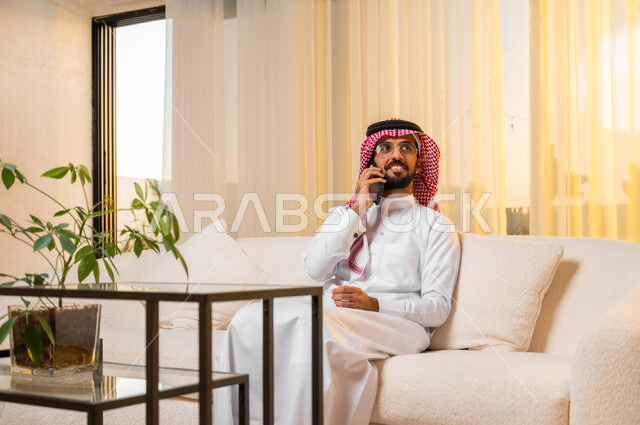 Communicating with friends and relatives using a mobile phone, answering calls and messages, hearing happy news via mobile phone, portrait of a young Saudi Arabian Gulf man wearing traditional clothing sitting on a comfortable sofa making a phone call, looking at something with gestures of joy and happiness