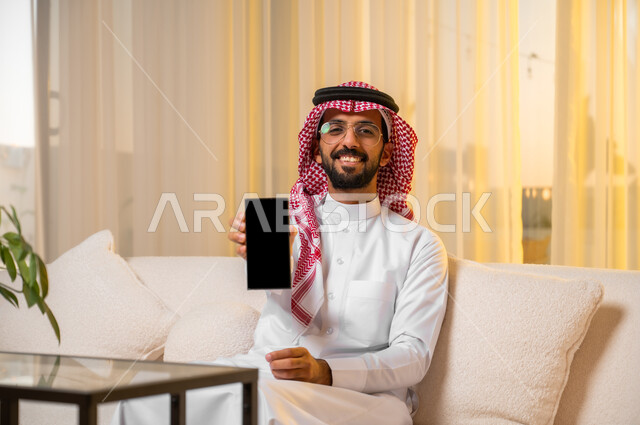 Mobile programs and applications, browsing social networking sites, the concept of the development of technology and modern techniques, a smiling young Saudi Arabian Gulf man wearing traditional clothing and glasses sitting on a comfortable sofa displaying a mobile phone with a blank black screen