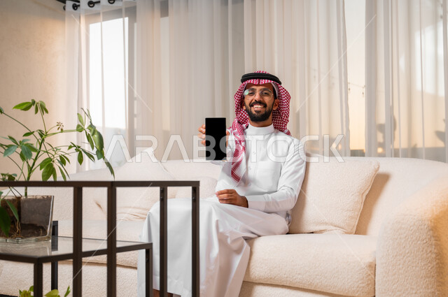 The concept of the development of technology and modern techniques, a smiling young Saudi Arabian Gulf man wearing traditional dress and glasses sitting on a comfortable sofa displaying a mobile phone with a blank black screen, mobile programs and applications, browsing social networking sites