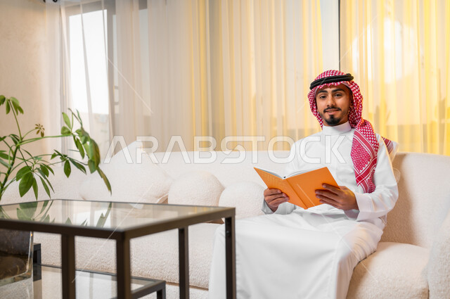 The concept of education and scientific research, spending free time learning and culture, a young Saudi Arabian Gulf man wearing traditional clothing sitting on a comfortable sofa reading a book, a hobby of reading and exploring, looking at the camera with gestures of joy and happiness