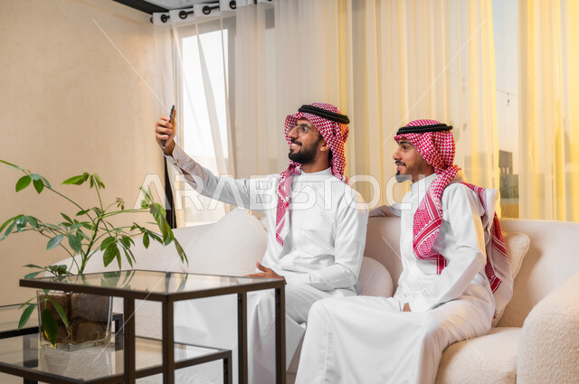 التقاط صورة سيلفي باستخدام الهاتف المحمول، شابان عربيان خليجيان سعوديان يرتديان الزي التقليدي يجلسان على أريكة مريحة يقومان باستعمال الأجهزة التقنية الحديثة في اخذ صور تذكارية، ايماءات وتعابير البهجة والسرور