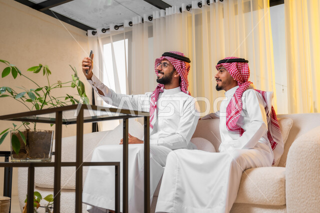 استعمال الأجهزة التقنية الحديثة في اخذ صور تذكارية، شابان عربيان خليجيان سعوديان يرتديات الزي التقليدي يجلسان على أريكة مريحة يقومان بإلتقاط صورة سيلفي باستخدام الهاتف المحمول، ايماءات وتعابير البهجة والسرور