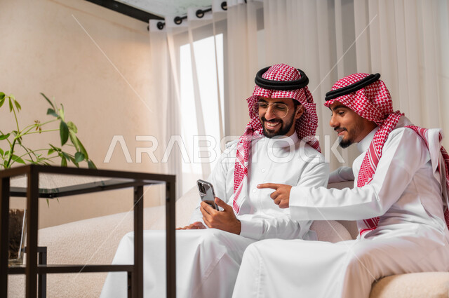 حوارات ونقاشات وجلسات شبابية، شابان عربيان خليجيان سعوديان يرتديات الزي التقليدي يجلسان على أريكة مريحة يقومان بإجراء مكالمة فيديو باستخدام الهاتف المحمول، استخدام الأجهزة التقنية الحديثة في التواصل مع الأصدقاء، ايماءات وتعابير البهجة والسرور