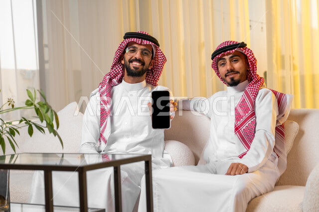النظر الى الكاميرا بإيماءات البهجة والسرور، شابان عربيان خليجيان سعوديان يرتديات الزي التقليدي يجلسان على أريكة مريحة يقومان بعرض شاشة سوداء فارغة باستخدام الهاتف المحمول، استخدام الأجهزة التقنية الحديثة