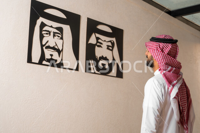 استذكار اليوم الوطني، مفهوم حب الوطن والانتماء اليه، صورة من الخلف لشاب عربي خليجي سعودي يرتدي الزي التقليدي يقف أمام لوحات فنية تحمل صور لملوك المملكة العربية السعودية، الفخر والاعتزاز بإنجازات البلاد