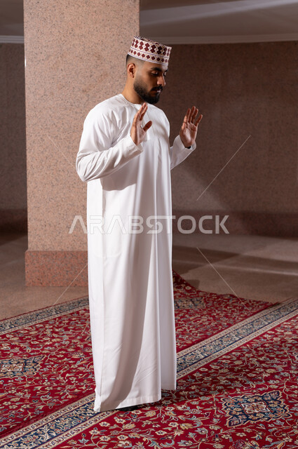 أداء الفرائض الدينية الاسلامية، مفهوم أركان الإسلام، رجل عربي خليجي عماني مسلم يرتدي الكمة والدشداشة يصلي داخل مصلى الشركة، العبادة والتقرب إلى الله، رفع اليدين للتكبير والشروع بالصلاة