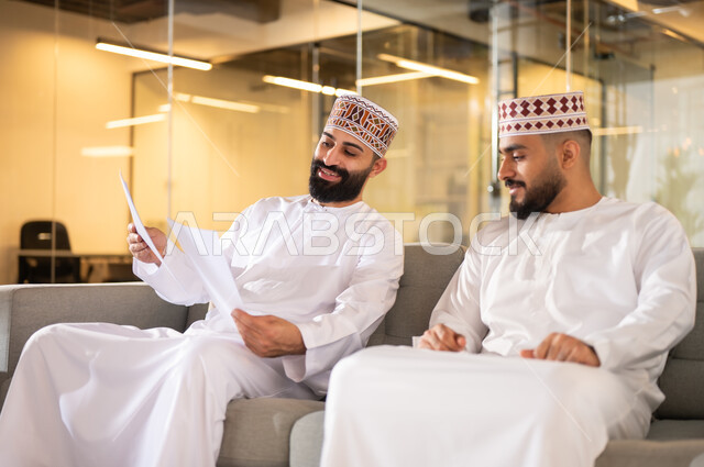 بيئة عمل مريحة، مهن ووظائف إدارية في سلطنة عمان، تبادل وجهات النظر بين الزملاء, إيماءات الهجة والسرور، موظفان عربيان خليجيان عمانيان يرتديان الدشداشة والكمة يجلسان على كنبة داخل مقر الشركة يقومان بالتعاون والتشارك في وضع الخطط