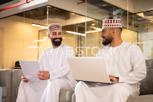Using technical devices to accomplish tasks, exchanging experiences and skills, the concept of partnership and cooperation, two Gulf Arab Omani employees wearing a dishdasha and a kummah sitting on a sofa inside the company headquarters using a laptop, administrative professions and jobs in the Sultanate of Oman, a comfortable work environment