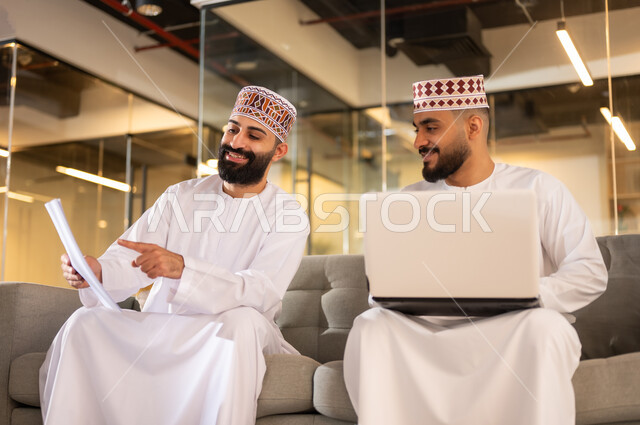 تبادل وجهات النظر بين الزملاء, إيماءات الهجة والسرور، التعاون والتشارك في وضع الخطط، موظفان عربيان خليجيان عمانيان يرتديان الدشداشة والكمة يجلسان على كنبة داخل مقر الشركة يقومان باستعمال الحاسوب المحمول، مهن ووظائف إدارية في سلطنة عمان، بيئة عمل مريحة