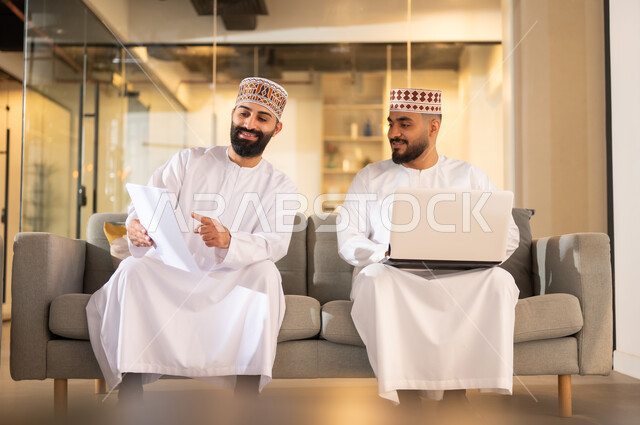 التعاون والتشارك في وضع الخطط والاستراتيجيات، تبادل الخبرات والمهارات, موظفان عربيان خليجيان عمانيان يرتديان الدشداشة والكمة يجلسان على كنبة داخل مقر الشركة يقومان باستعمال الحاسوب المحمول، مهن ووظائف إدارية في سلطنة عمان، بيئة عمل مريحة