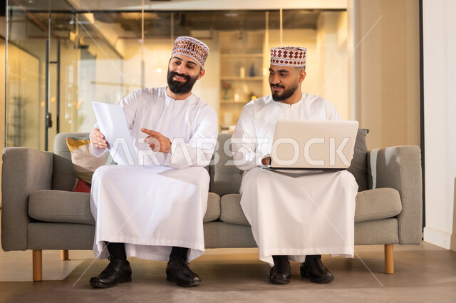 تبادل وجهات النظر مع الزملاء, مفهوم الشراكة والتعاون, استخدام الأجهزة التقنية الحديثة، موظفان عربيان خليجيان عمانيان يرتديان الدشداشة والكمة يجلسان داخل مقر الشركة يقومان باستعمال الحاسوب المحمول في إنجاز المهام، مهن ووظائف إدارية في سلطنة عمان، بيئة عمل مريحة