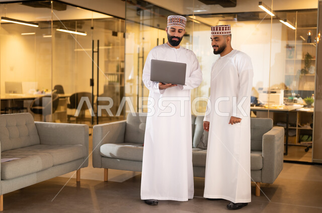 استخدام الأجهزة التقنية الحديثة، تبادل وجهات النظر مع الزملاء, مفهوم الشراكة والتعاون, موظفان عربيان خليجيان عمانيان يرتديان الدشداشة والكمة يقفان داخل مقر الشركة يقومان باستعمال الحاسوب المحمول في إنجاز المهام، مهن ووظائف إدارية في سلطنة عمان، بيئة عمل مريحة