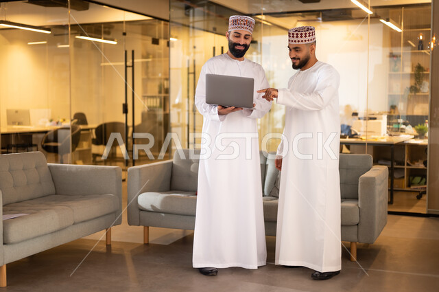 تبادل وجهات النظر مع الزملاء, مفهوم الشراكة والتعاون, استخدام الأجهزة التقنية الحديثة، موظفان عربيان خليجيان عمانيان يرتديان الدشداشة والكمة يقفان داخل مقر الشركة يقومان باستعمال الحاسوب المحمول في إنجاز المهام، مهن ووظائف إدارية في سلطنة عمان، بيئة عمل مريحة