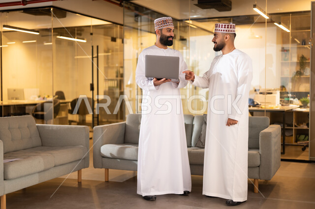 مفهوم الشراكة والتعاون, تبادل وجهات النظر مع الزملاء, استخدام الأجهزة التقنية الحديثة، موظفان عربيان خليجيان عمانيان يرتديان الدشداشة والكمة يقفان داخل مقر الشركة يقومان باستعمال الحاسوب المحمول في إنجاز المهام، مهن ووظائف إدارية في سلطنة عمان، بيئة عمل مريحة