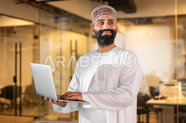 Using modern technical devices to accomplish tasks, gestures of joy and happiness, administrative professions and jobs in the Sultanate of Oman, a comfortable environment in Omani companies, an Arab Gulf Omani employee wearing a dishdasha and a kuma standing inside the company headquarters clicking on the laptop