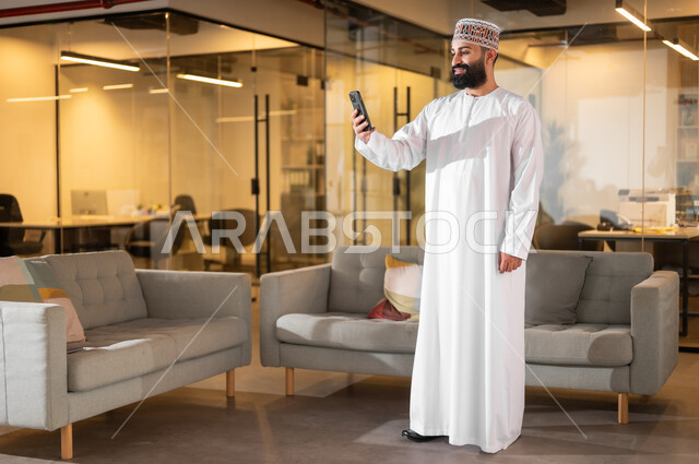 بيئة مريحة في الشركات العمانية، مهن ووظائف إدارية في سلطنة عمان، الوقوف باستقامة والنظر إلى الجوال بإيماءات البهجة والسرور، موظف عربي خليجي عماني يرتدي الدشداشة والكمة يقوم باستخدام الهاتف المحمول داخل مقر الشركة