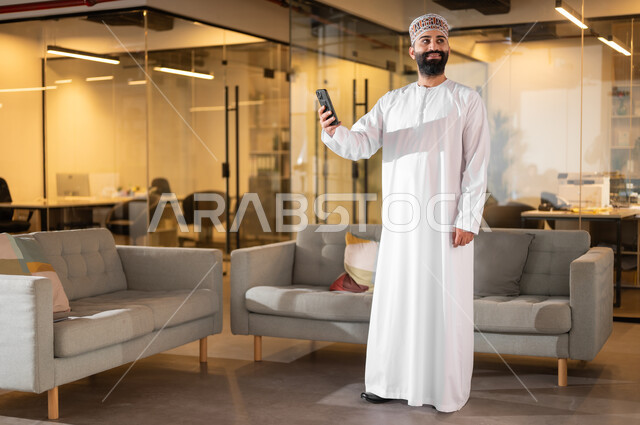 بيئة مريحة في الشركات العمانية، مهن ووظائف إدارية في سلطنة عمان، الوقوف باستقامة والنظر إلى شيء ما بإيماءات البهجة والسرور، موظف عربي خليجي عماني يرتدي الدشداشة والكمة يقوم باستخدام الهاتف المحمول داخل مقر الشركة