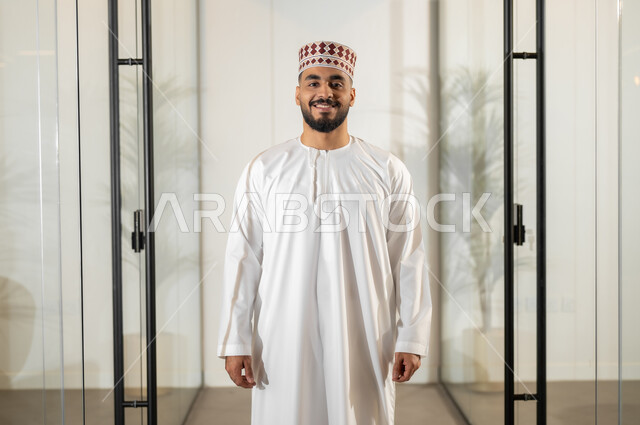 الوقوف باستقامة والنظر إلى الكاميرا بإيماءات البهجة والسرور، بيئة مريحة في الشركات العمانية، مهن ووظائف إدارية في سلطنة عمان، موظف عربي خليجي عماني يرتدي الدشداشة والكمة يقف داخل مقر الشركة