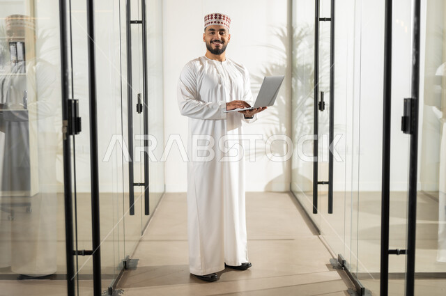 Using modern technical devices to accomplish tasks, gestures of joy and happiness, administrative professions and jobs in the Sultanate of Oman, a comfortable environment in Omani companies, an Arab Gulf Omani employee wearing a dishdasha and a kuma standing inside the company headquarters clicking on the laptop