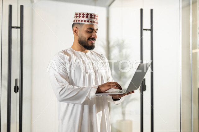 بيئة مريحة في الشركات العمانية، استخدام الأجهزة التقنية الحديثة في إنجاز المهام، إيماءات البهجة والسرور، مهن ووظائف إدارية في سلطنة عمان، موظف عربي خليجي عماني يرتدي الدشداشة والكمة يقف داخل مقر الشركة يقوم بالنقر على الحاسوب المحمول