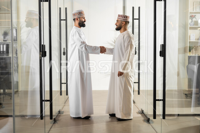 إيماءات البهجة والسرور، التعرف إلى الزملاء, مفهوم الشراكة والتعاون, صورة من الجانب لموظفان عربيان خليجيان عمانيان يرتديان الدشداشة والكمة يقفان داخل مقر الشركة يقومان بمصافحة بعضهما البعض، مهن ووظائف إدارية في سلطنة عمان، بيئة عمل مريحة