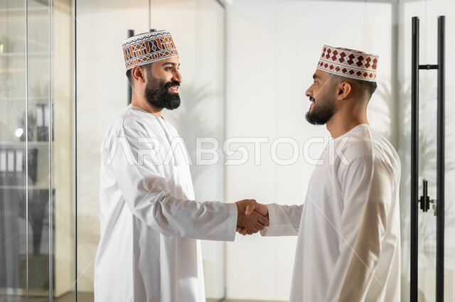 التعرف إلى الزملاء وإلقاء التحية عليهم, مفهوم الشراكة والتعاون, إيماءات البهجة والسرور، صورة من الجانب لموظفان عربيان خليجيان عمانيان يرتديان الدشداشة والكمة يقفان داخل مقر الشركة يقومان بمصافحة بعضهما البعض، مهن ووظائف إدارية في سلطنة عمان، بيئة عمل مريحة