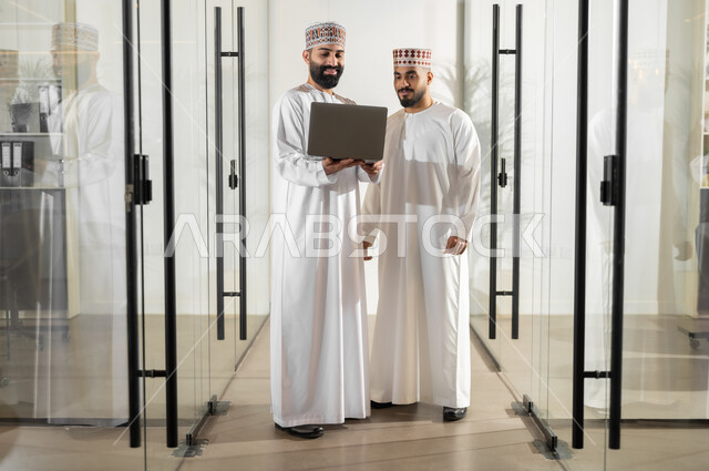 مفهوم الشراكة والتعاون, تبادل وجهات النظر مع الزملاء, استخدام الأجهزة التقنية الحديثة، موظفان عربيان خليجيان عمانيان يرتديان الدشداشة والكمة يقفان داخل مقر الشركة يقومان باستعمال الحاسوب المحمول في إنجاز المهام، مهن ووظائف إدارية في سلطنة عمان، بيئة عمل مريحة