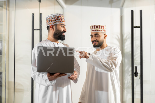 تبادل وجهات النظر مع الزملاء, استخدام الأجهزة التقنية الحديثة، مفهوم الشراكة والتعاون, موظفان عربيان خليجيان عمانيان يرتديان الدشداشة والكمة يقفان داخل مقر الشركة يقومان باستعمال الحاسوب المحمول في إنجاز المهام، مهن ووظائف إدارية في سلطنة عمان، بيئة عمل مريحة