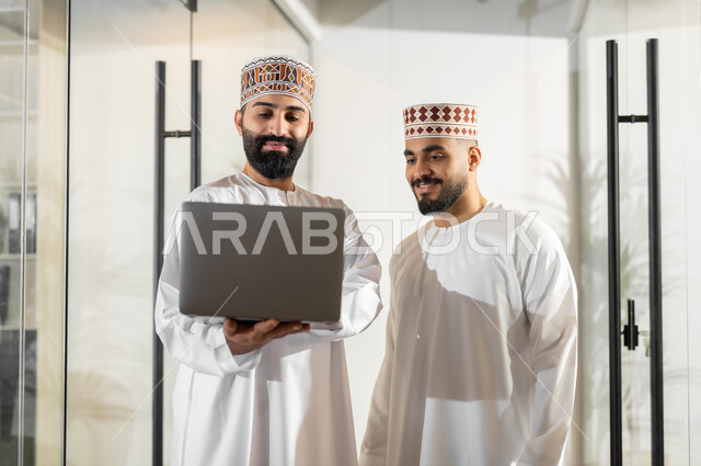 استخدام الأجهزة التقنية الحديثة في إنجاز المهام، تبادل الخبرات و المهارات, مفهوم الشراكة والتعاون, موظفان عربيان خليجيان عمانيان يرتديان الدشداشة والكمة يقفان داخل مقر الشركة يقومان باستعمال الحاسوب المحمول، مهن ووظائف إدارية في سلطنة عمان، بيئة عمل مريحة