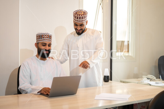 مهن ووظائف إدارية في سلطنة عمان، مفهوم الشراكة والتعاون, تبادل الخبرات و المهارات, استخدام الأجهزة التقنية الحديثة في إنجاز المهام، موظفان عربيان خليجيان عمانيان يرتديان الدشداشة والكمة يجلسان داخل مكتب يقومان باستعمال الحاسوب المحمول، بيئة عمل مريحة