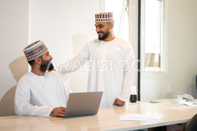 مفهوم الشراكة والتعاون, تبادل الخبرات و المهارات, استخدام الأجهزة التقنية الحديثة في إنجاز المهام، موظفان عربيان خليجيان عمانيان يرتديان الدشداشة والكمة يجلسان داخل مكتب يقومان باستعمال الحاسوب المحمول، مهن ووظائف إدارية في سلطنة عمان، بيئة عمل مريحة
