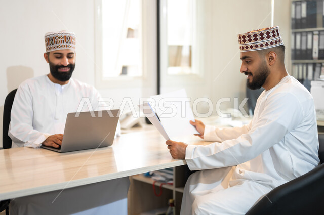 تبادل الخبرات و المهارات, استخدام الأجهزة التقنية الحديثة في إنجاز المهام، مفهوم الشراكة والتعاون, موظفان عربيان خليجيان عمانيان يرتديان الدشداشة والكمة يجلسان داخل مكتب يقومان باستعمال الحاسوب المحمول، مهن ووظائف إدارية في سلطنة عمان، بيئة عمل مريحة