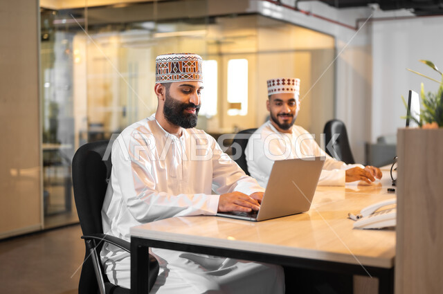 Using modern technical devices, gestures of joy and happiness, cooperation and sharing in performing tasks, two Gulf Arab Omani employees wearing a dishdasha and a cloak sitting in an office using a laptop, administrative professions and jobs in the Sultanate of Oman, a comfortable work environment