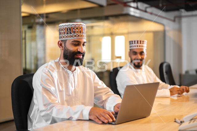 ايماءات البهجة والسرور، التعاون التشارك في أداء المهام، استخدام الأجهزة التقنية الحديثة، موظفان عربيان خليجيان عمانيان يرتديان الدشداشة والكمة يجلسان داخل مكتب يقومان باستعمال الحاسوب المحمول، مهن ووظائف إدارية في سلطنة عمان، بيئة عمل مريحة