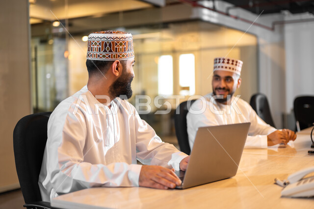 تبادل الأحديث ضمن مقر الشركة، التعاون التشارك في أداء المهام، استخدام الأجهزة التقنية الحديثة، موظفان عربيان خليجيان عمانيان يرتديان الدشداشة والكمة يجلسان داخل مكتب يقومان باستعمال الحاسوب المحمول، مهن ووظائف إدارية في سلطنة عمان، بيئة عمل مريحة